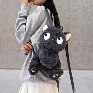 Może przedstawiać: Pluszowy, ciemnoszary plecak w kształcie kota z dużymi białymi oczami, różowym nosem i podkładkami na łapy. Plecak ma czarne paski i puszystą fakturę, przypominającą pluszową zabawkę. Uszy kota są jasno różowe.