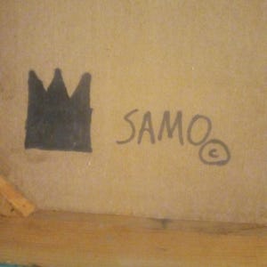 Puede incluir: Una corona negra y la palabra "SAMO" con un s&iacute;mbolo de copyright escrito en tinta negra sobre una superficie marr&oacute;n.