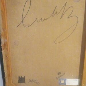 Puede incluir: Un primer plano de la parte posterior de una pintura enmarcada. La pintura est&aacute; firmada "SAMO" y "Basquiat" con marcador negro. El marco es de madera y tiene un acabado marr&oacute;n.