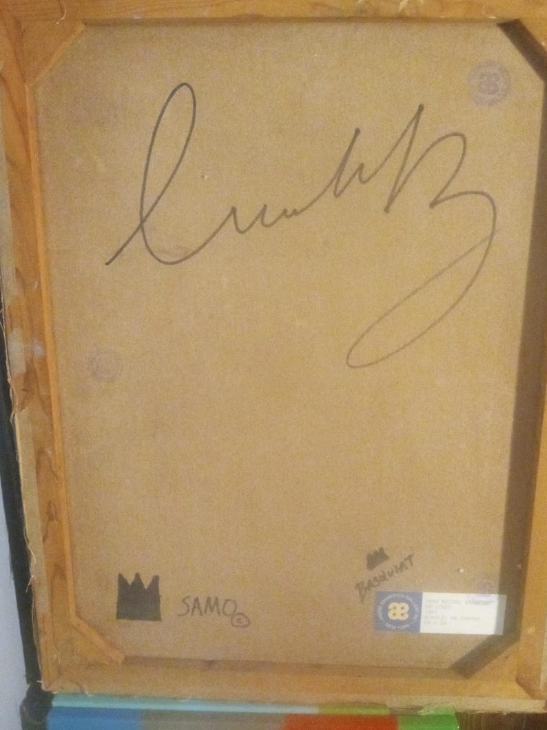 Puede incluir: Un primer plano de la parte posterior de una pintura enmarcada. La pintura est&aacute; firmada "SAMO" y "Basquiat" con marcador negro. El marco es de madera y tiene un acabado marr&oacute;n.