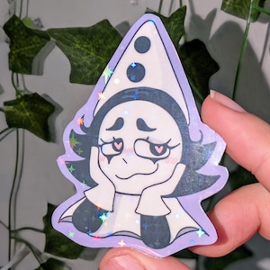 May include: A holographic sticker featuring a cartoon character with dark hair and a white pointed hat with two black circles. The character has heart-shaped pupils, rosy cheeks, and is resting their chin on their hands with a light purple border.