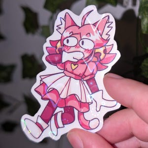 May include: A holographic sticker featuring a cartoon character with pink hair, cat ears, and a tail. The character wears a pink top with a yellow heart, a white skirt, and white gloves. The design has a glossy, reflective finish with heart-shaped light refractions.