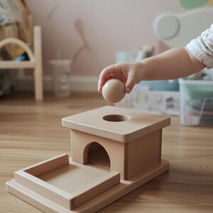 Könnte beinhalten: Ein Holzspielzeug für die frühe Kindheitsentwicklung. Es hat eine quadratische Oberseite mit einem Loch, eine bogenförmige Öffnung und eine Ablage. Eine kleine Holzkugel wird über dem Loch gehalten. Die Maße sind in Zentimetern.