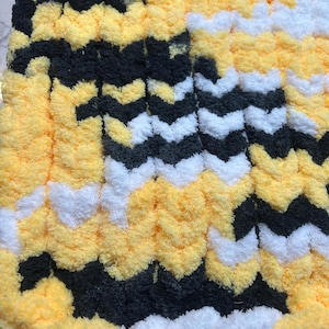 May include: A close-up of a soft, textured blanket with a chevron pattern. The blanket features alternating rows of yellow, black, and white. The plush material creates a cozy and inviting appearance, perfect for home decor.