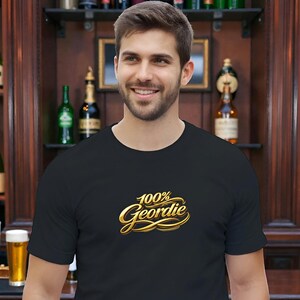 K&ouml;nnte beinhalten: Schwarzes T-Shirt mit Rundhalsausschnitt und dem goldenen Schriftzug "100% Geordie". Die Abbildung zeigt eine Person in einer Bar, mit Regalen voller Flaschen im Hintergrund.