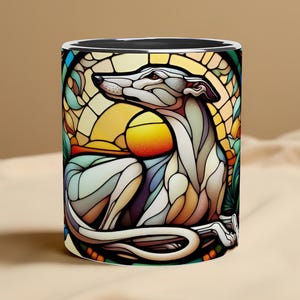 May include: A ceramic mug with a stained-glass style design of a greyhound dog. The artwork includes a yellow sun and the dog is rendered in white, grey, and brown. The mug has a black interior and a white rim, suitable for hot beverages.