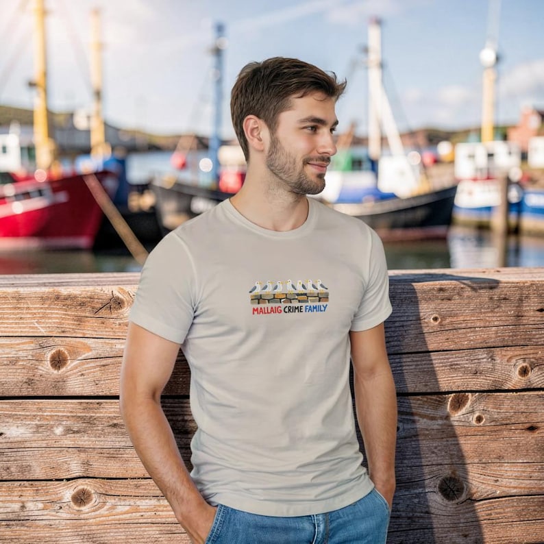 May include: A light grey t-shirt with a graphic of seagulls and the text "Mallaig Crime Family" in red, blue, and yellow. The person is standing near a wooden dock with boats in the background.