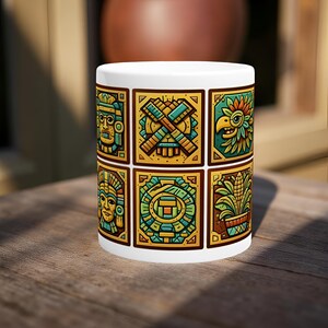 May include: White ceramic mug featuring six colorful Aztec-inspired designs in gold, teal, and brown. The designs include stylized faces, crossed tools, a bird, and geometric patterns. The mug is sitting on a wooden surface.