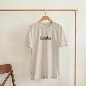 May include: A cream-coloured t-shirt with a crew neck and short sleeves. The shirt displays a graphic of birds on a ledge, with the text "MALLAIG CRIME FAMILY" underneath. The t-shirt is hanging on a wooden hanger.