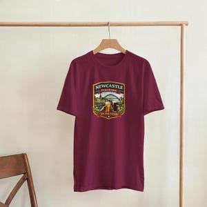 Pu&ograve; includere: T-shirt bordeaux con una grafica con la scritta "Newcastle Pub Crawl" e un'illustrazione di un ponte, edifici e birra. Il design &egrave; a forma di scudo con un bordo dorato. La maglietta &egrave; appesa a un appendiabiti di legno.