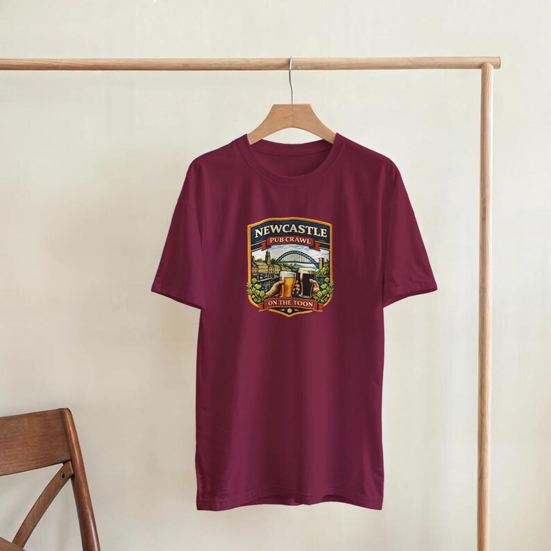 Pu&ograve; includere: T-shirt bordeaux con una grafica con la scritta "Newcastle Pub Crawl" e un'illustrazione di un ponte, edifici e birra. Il design &egrave; a forma di scudo con un bordo dorato. La maglietta &egrave; appesa a un appendiabiti di legno.