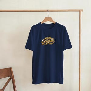 K&ouml;nnte beinhalten: Ein marineblaues T-Shirt mit dem Text "100% Geordie" in goldener Schrift. Das T-Shirt h&auml;ngt an einer h&ouml;lzernen Kleiderstange. Das Shirt hat kurze &Auml;rmel und einen Rundhalsausschnitt.