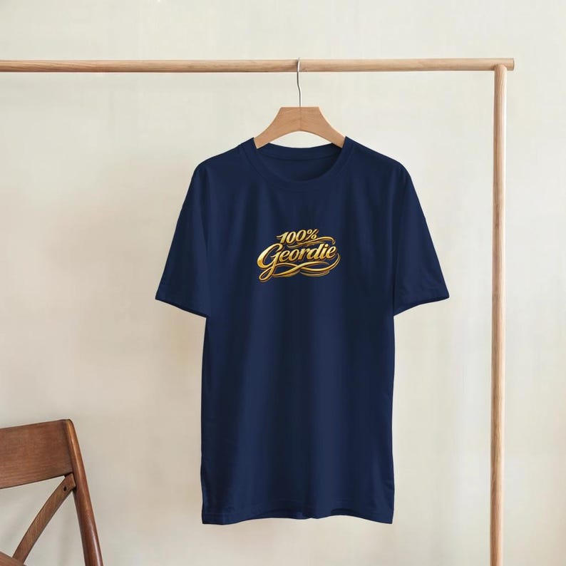 K&ouml;nnte beinhalten: Ein marineblaues T-Shirt mit dem Text "100% Geordie" in goldener Schrift. Das T-Shirt h&auml;ngt an einer h&ouml;lzernen Kleiderstange. Das Shirt hat kurze &Auml;rmel und einen Rundhalsausschnitt.