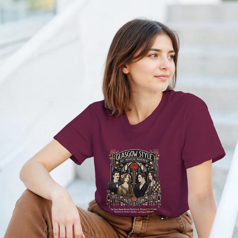 Puede incluir: Camiseta burdeos con un dise&ntilde;o gr&aacute;fico del movimiento Art Nouveau de Glasgow. El dise&ntilde;o incluye el texto "Glasgow Style Art Nouveau Movement" e ilustraciones de personas. La camiseta es de cuello redondo y manga corta.