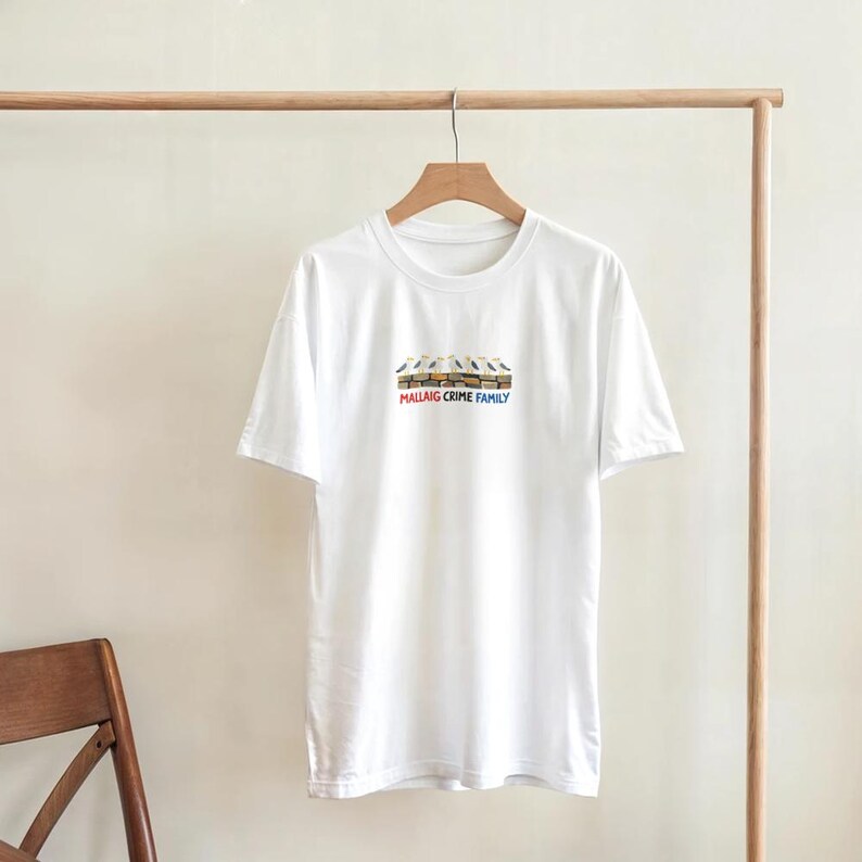 May include: A white t-shirt with a crew neck and short sleeves. The shirt displays a graphic of birds on a ledge, with the text "MALLAIG CRIME FAMILY" beneath. The t-shirt is suspended from a wooden hanger on a clothes rail.
