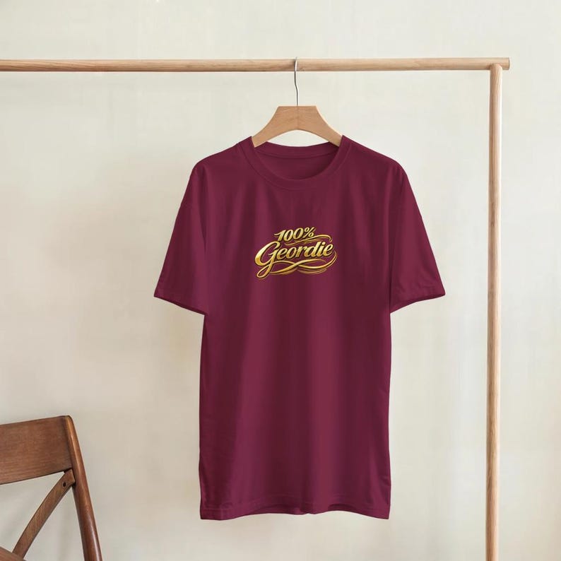 K&ouml;nnte beinhalten: Burgunderfarbenes T-Shirt mit dem Text "100% Geordie" in goldener Schrift. Das T-Shirt h&auml;ngt an einem Holzkleiderb&uuml;gel. Das T-Shirt hat kurze &Auml;rmel und einen Rundhalsausschnitt. Der Hintergrund ist neutral.