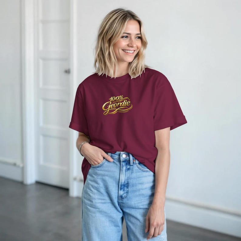K&ouml;nnte beinhalten: Ein weinrotes T-Shirt mit dem Text "100% Geordie" in goldener Schrift. Das Shirt hat einen Rundhalsausschnitt und wird mit einer Jeans getragen. Das Shirt ist aus einem weichen Material.