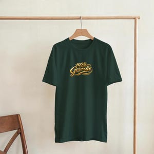 K&ouml;nnte beinhalten: Ein dunkelgr&uuml;nes T-Shirt mit dem Text "100% Geordie" in goldener Schrift. Das Shirt h&auml;ngt an einer h&ouml;lzernen Kleiderstange. Der Text ist in einer stilisierten Schriftart.