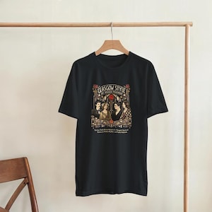 Puede incluir: Camiseta negra con un dise&ntilde;o gr&aacute;fico. El dise&ntilde;o presenta el texto "GLASGOW STYLE ART NOUVEAU MOVEMENT" e ilustraciones de cuatro figuras y motivos florales en oro, rojo y marr&oacute;n. La camiseta est&aacute; colgada en una percha de madera.