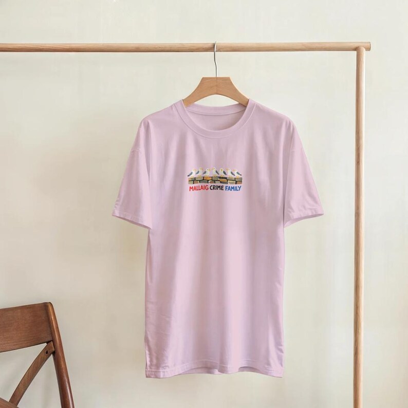 May include: A pale pink t-shirt with a crew neck and short sleeves. The shirt displays a graphic of seagulls above the text "Mallaig Crime Family" in red, white, and blue.