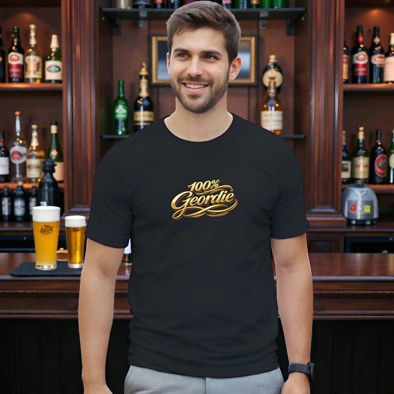 K&ouml;nnte beinhalten: Schwarzes T-Shirt mit Rundhalsausschnitt und dem goldenen Schriftzug "100% Geordie". Die Abbildung zeigt eine Person in einer Bar, mit Regalen voller Flaschen im Hintergrund.
