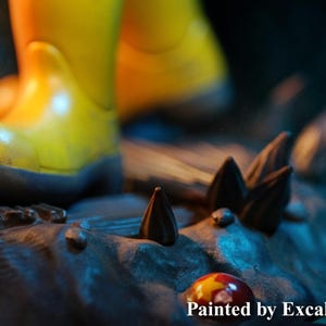 May include: A close-up of a figurine with yellow boots standing on a rocky surface with dark brown spikes. The base is painted in shades of gray and blue. The text "Painted by ExcaliburCreations" is visible at the bottom.