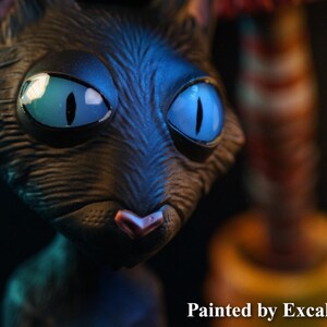 May include: A close-up of a black cat sculpture with large, bright blue eyes and a pink nose. The sculpture is painted by ExcaliburCreations. The cat's fur appears textured, and the background is dark, highlighting the cat's features.