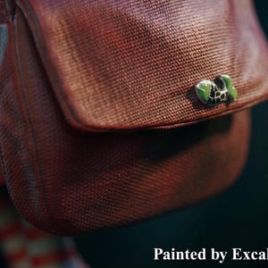 May include: Close-up of a brown satchel with a flap and a green and black decorative clasp. The satchel has a textured appearance. The text "Painted by ExcaliburCreations" is at the bottom.