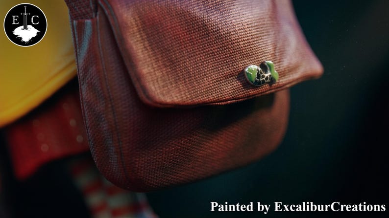 May include: Close-up of a brown satchel with a flap and a green and black decorative clasp. The satchel has a textured appearance. The text "Painted by ExcaliburCreations" is at the bottom.