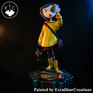 May include: A figurine of a girl in a yellow raincoat and boots, holding a green marble eye. A black cat with blue eyes sits beside her. The base is striped with a figure lying down. The text "Painted by ExcaliburCreations" is at the bottom.