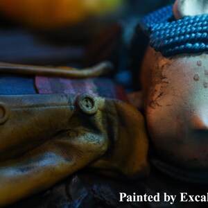 May include: A detailed close-up of a doll with a pale face, button eyes, and blue braided hair. The doll wears a yellow coat with brown buttons. The text "Painted by ExcaliburCreations" is visible at the bottom.