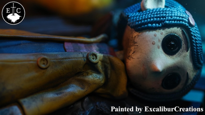 May include: A detailed close-up of a doll with a pale face, button eyes, and blue braided hair. The doll wears a yellow coat with brown buttons. The text "Painted by ExcaliburCreations" is visible at the bottom.