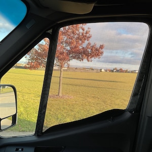 May include: A black mesh window screen is installed in a car window. The screen is partially open, revealing a view of a grassy field with a red-leaved tree in the foreground. The sky is blue with white clouds.