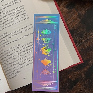 Puede incluir: Un marcapáginas holográfico con un degradado de azul a morado. El marcapáginas presenta una serie de coloridas polillas y estrellas. La parte superior e inferior del marcapáginas tienen un borde decorativo con lunas crecientes.