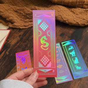May include: A hand holds a red holographic bookmark with a dragon design, suns, and geometric patterns. Other bookmarks with similar designs are visible. The bookmarks have a shiny, iridescent finish, and are likely made of paper or cardstock.