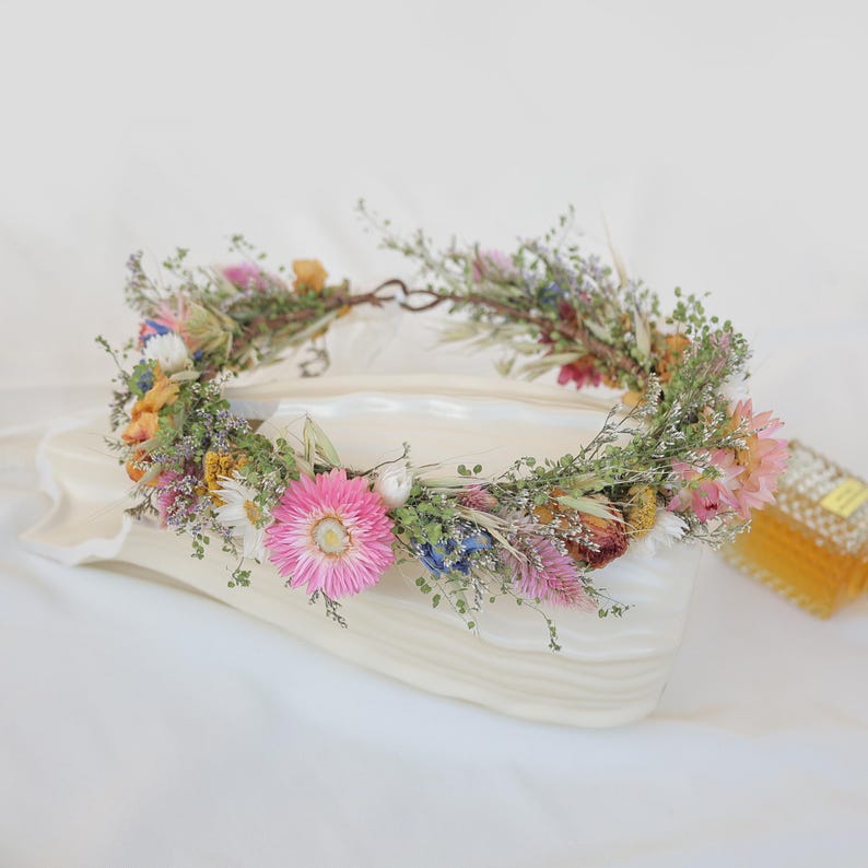 May include: A floral crown featuring a variety of dried flowers in shades of pink, yellow, blue, and green. The crown is made of a natural vine base and is ideal for weddings or special events.