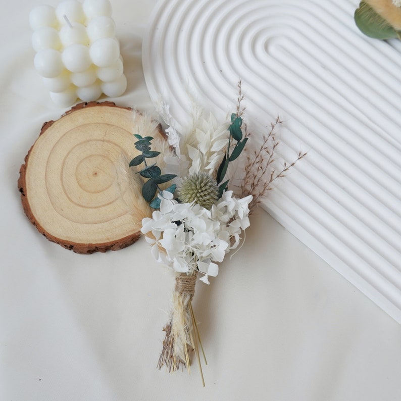 May include: A small bouquet of dried flowers in shades of white and green, tied with twine. The bouquet includes white hydrangeas, green eucalyptus leaves, and dried grasses.