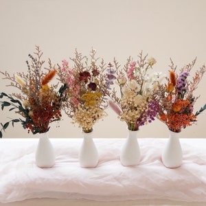 May include: Five small white vases filled with dried flower arrangements. The arrangements feature a variety of colours and textures, including pink, purple, white, orange, and brown.