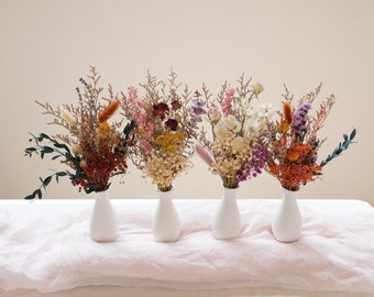Op de afbeelding: Vijf kleine witte vazen gevuld met gedroogde bloemenarrangementen. De arrangementen bevatten een verscheidenheid aan kleuren en texturen, waaronder roze, paars, wit, oranje en bruin.