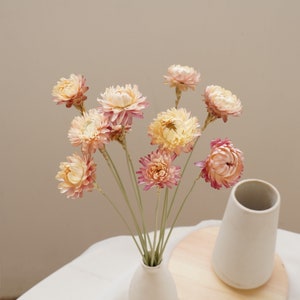 May include: A bouquet of dried flowers in shades of pink and cream, arranged in a white vase. The flowers have a delicate, papery texture and are a popular choice for home decor.