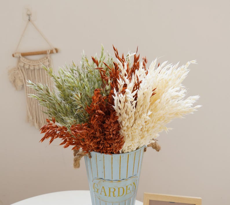 May include: A bouquet of dried grasses in a blue and white striped metal vase with the word "GARDEN" printed on the side. The bouquet includes beige, brown, and green dried grasses.