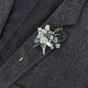 Puede incluir: Un prendedor floral con flores y follaje blancos y verdes, prendido a una blazer de tweed gris oscuro. El prendedor incluye peque&ntilde;as flores blancas, hojas verdes y una pieza blanca. Tambi&eacute;n es visible un alfiler dorado.
