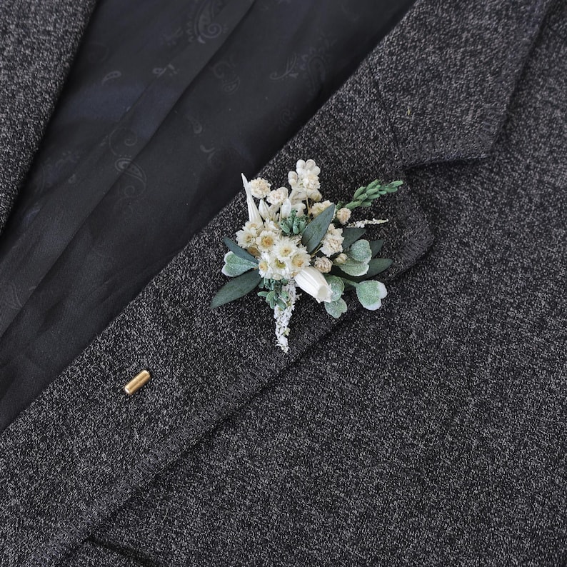 Puede incluir: Un prendedor floral con flores y follaje blancos y verdes, prendido a una blazer de tweed gris oscuro. El prendedor incluye peque&ntilde;as flores blancas, hojas verdes y una pieza blanca. Tambi&eacute;n es visible un alfiler dorado.