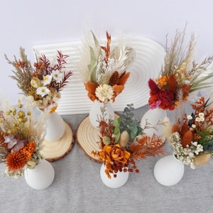 May include: Six small white vases filled with dried flower arrangements in shades of brown, orange, white, and red. The vases are arranged on a white surface with wooden rounds underneath.