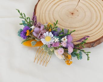 May include: A floral hair comb featuring a variety of dried flowers in shades of purple, yellow, white, and green. The flowers are arranged on a gold-colored comb, perfect for weddings or special occasions.