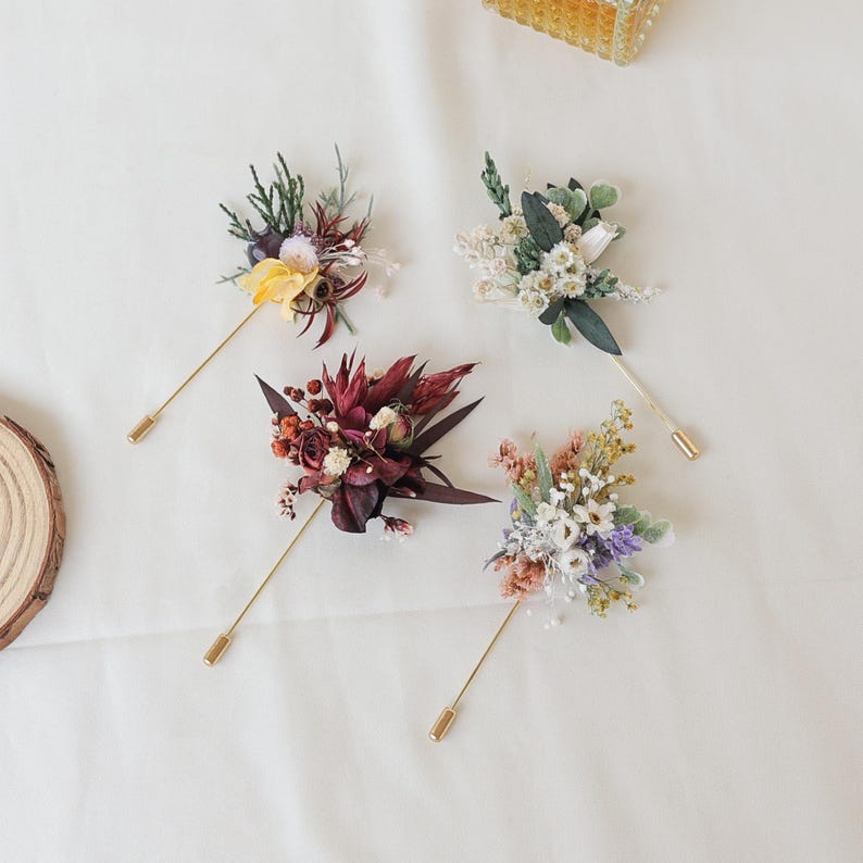 Puede incluir: Cuatro prendedores florales con tallos dorados y arreglos florales decorativos. Las flores incluyen una variedad de colores, como amarillo, blanco, morado y burdeos. Los prendedores est&aacute;n dispuestos sobre una superficie blanca.