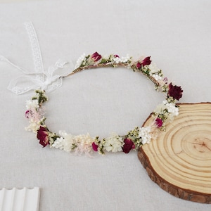 May include: A delicate floral crown featuring white and burgundy flowers, tied with a white ribbon. The crown is made from dried flowers and is ideal for a wedding or special occasion.