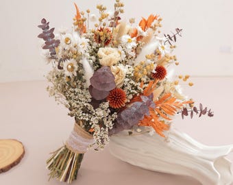 May include: A dried flower bouquet featuring white daisies, orange plumes, purple eucalyptus leaves, and small yellow buds. The stems are bound together with a tan burlap ribbon and a strip of white lace, resting beside a white ceramic vase.