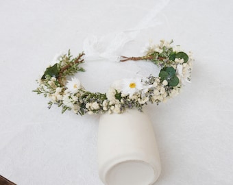 May include: A floral crown featuring white daisy-like flowers, small white baby's breath, green eucalyptus leaves, and tiny purple filler flowers. The circular arrangement is constructed on a brown vine base with a white lace ribbon tie at the back.