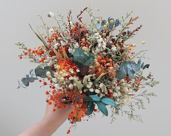 Peut inclure: Un bouquet de fleurs séchées composé d'un mélange de fleurs orange, blanches et rouge foncé, agrémenté de feuilles d'eucalyptus vertes et de délicates tiges de gypsophile. L'arrangement inclut diverses textures d'herbes séchées et de fleurs.